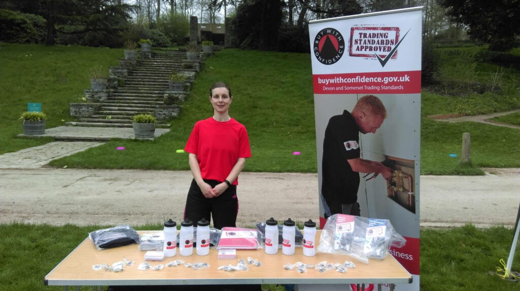 Rachael promoting Buy With Confidence - she stands behind a table covered with Buy With Confidence water bottles and other merchandise. Next to her is a stand-up banner.